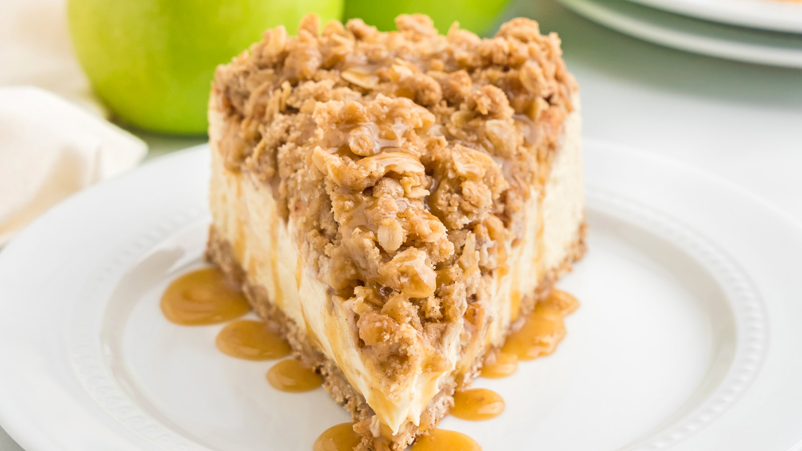 Slice of apple crumble cheesecake on a white plate and drizzled with caramel sauce.