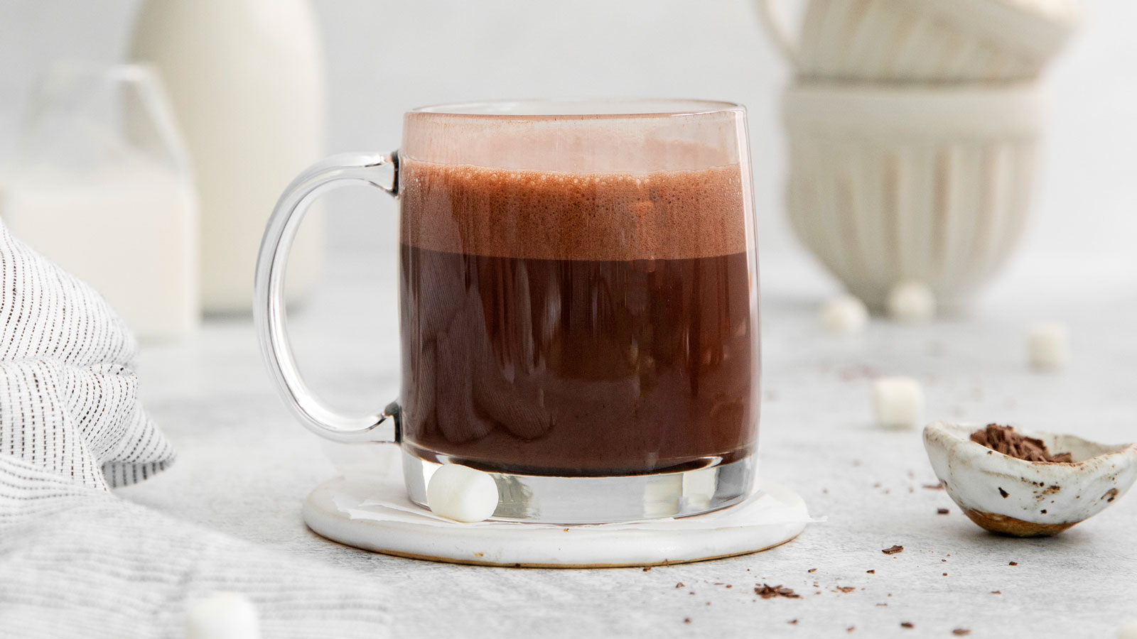 Clear mug with hot chocolate made with frothed milk with a milk bottle and more cups in the background.