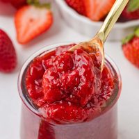 Close-up photo of a glass jar of homemade strawberry preserves with a golden spoon in the background you see fresh strawberries scattered about.