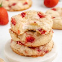 Photo of three strawberry cheesecake cookies stacked on each other with the top on with a bite taken out showing the creamy cheesecake filling.