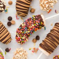 overhead shot of several colorful banana pops decorated with different toppings and strips with scattered chocolate and peanut butter chips on a white background