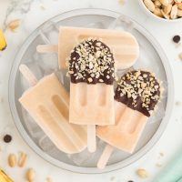 Overhead photo showing creamy banana popsicles on a plate, two of which have been dipped in chocolate and rolled in chopped peanuts.