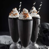 Photo of three black milkshakes topped with whipped cream and orange and black sprinkles to show how these milkshakes are perfect for Halloween. The milkshake is super black due to black cocoa and has a taste like oreo's.