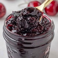 Photo of a jar of cherry preserves with a spoon lifting the homemade preserves out of the jar