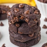 Photo of a stack of 4 banana cocoa cookies that look rich and chocolaty loaded with tons of chocolate chips and in the background you see a bright yellow banana