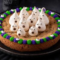 Photo of a Halloween cookie cake with green and purple rim with candy eyeballs for decoration in the middle of the cake are several meringue ghosts making it a perfect Halloween dessert